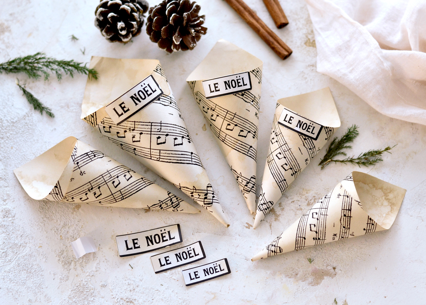 Five sheet music paper cones for Christmas laying on the table with two pine cones, 2 cinnamon sticks, greenery and small 'LE NOEL' tabs