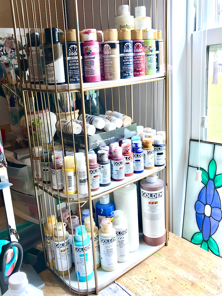 3 tier shelf stores acrylic paint bottles on work table make for easy Craft Room Storage Ideas on a Budget